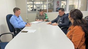 Três homens e uma mulher sentados em uma mesa de reunião. Homem com farda da Brigada Militar assina um papel